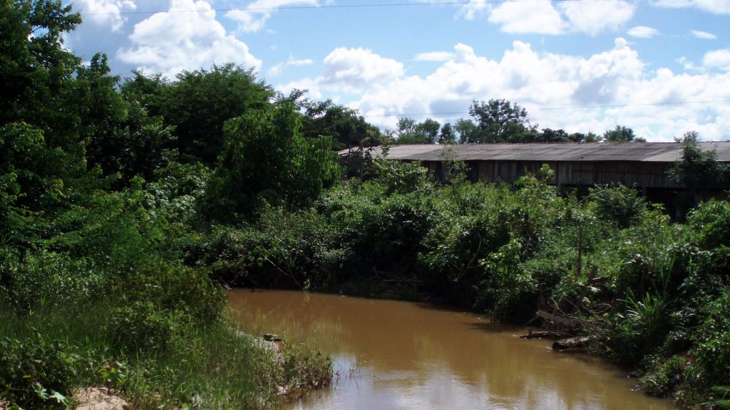 Ein kleiner Fluss außerhalb von Juara. Im Hintergrund ein landwirtschaftliches Gebäude aus Holz.